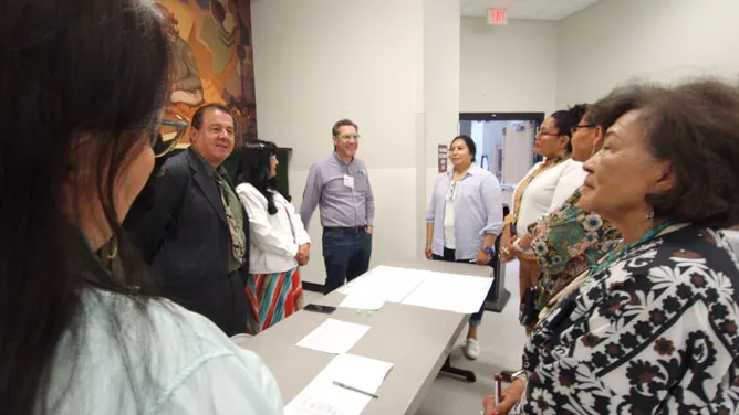 A group of adults standing in a semi-circle around a table with papers on it, engaged in conversation.