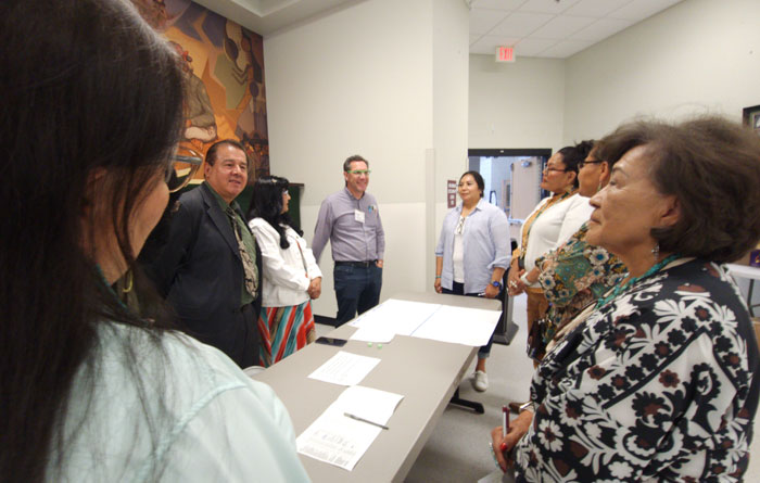 A group of adults standing in a semi-circle around a table with papers on it, engaged in conversation.
